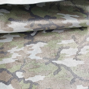 May include: Close-up of a camouflage pattern fabric in shades of green, brown, and gray. The fabric has a textured surface and a folded edge, revealing a lighter underside. The pattern is a blend of organic shapes.
