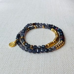 May include: A double-strand bracelet with dark blue gemstone beads and gold accents. The bracelet features a small gold charm with a design.