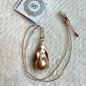 May include: A gold-toned necklace with a large, irregularly shaped, iridescent pearl pendant. The pendant is a creamy, pearlescent color with hints of pink and peach. A white tag with the text "soul star meditation" is attached.