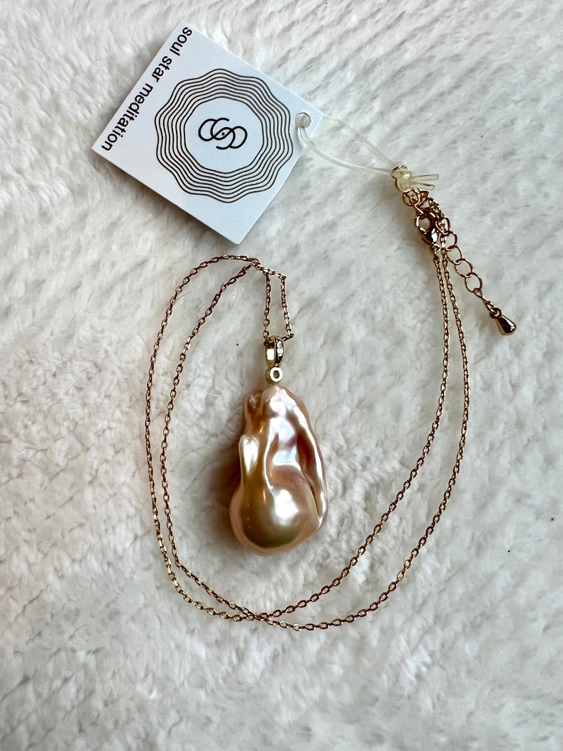May include: A gold-toned necklace with a large, irregularly shaped, iridescent pearl pendant. The pendant is a creamy, pearlescent color with hints of pink and peach. A white tag with the text "soul star meditation" is attached.