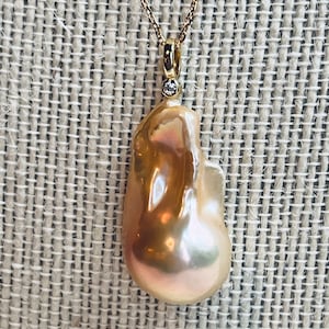 May include: A gold necklace featuring a large, irregularly shaped, lustrous pearl pendant. The pearl has a warm, peachy-pink hue with iridescent highlights. A small diamond accent is set above the pearl. The chain is delicate and gold.