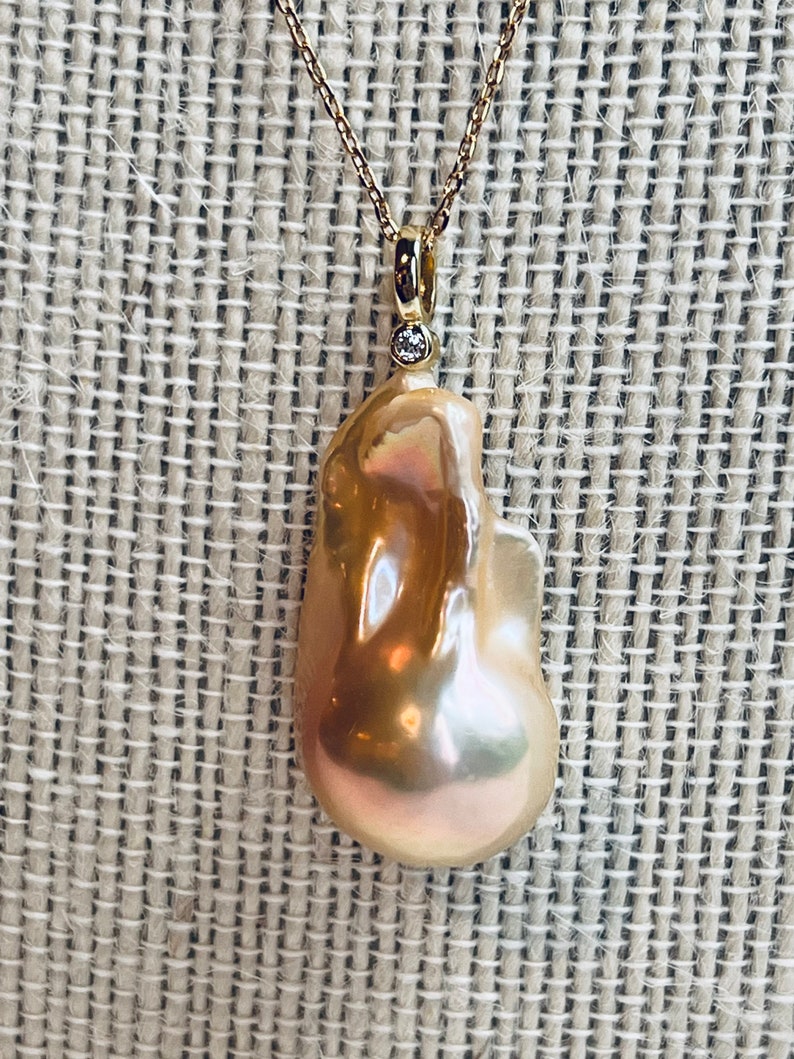 May include: A gold necklace featuring a large, irregularly shaped, lustrous pearl pendant. The pearl has a warm, peachy-pink hue with iridescent highlights. A small diamond accent is set above the pearl. The chain is delicate and gold.