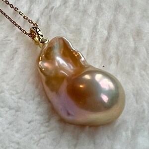 May include: A gold-toned necklace featuring a large, irregularly shaped pearl pendant. The pearl has a lustrous, iridescent surface with hues of pink, gold, and cream. The chain is delicate and appears to be gold. The pendant is the focal point.