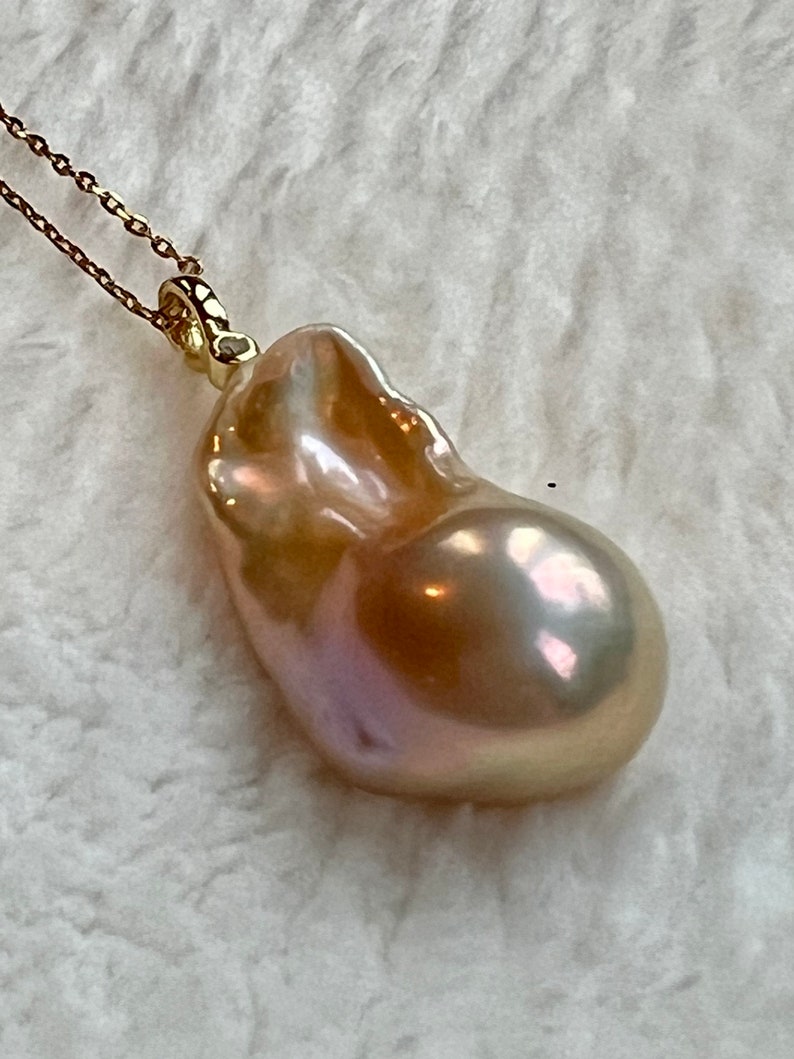 May include: A gold-toned necklace featuring a large, irregularly shaped pearl pendant. The pearl has a lustrous, iridescent surface with hues of pink, gold, and cream. The chain is delicate and appears to be gold. The pendant is the focal point.