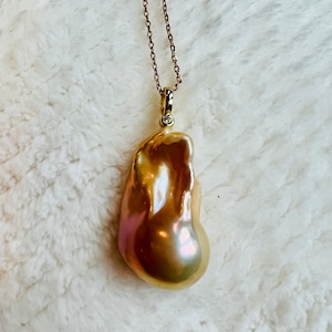 May include: A gold-toned necklace featuring a large, irregularly shaped pearl pendant. The pearl has a lustrous, iridescent surface with hues of gold, pink, and purple. A small diamond accents the pendant's bail. The chain is delicate and gold-colored.