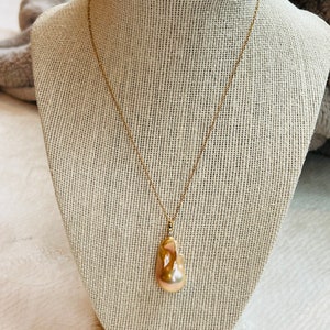 May include: A gold-colored necklace with a large, irregularly shaped, peach-colored pearl pendant. The pearl hangs from a delicate gold chain, displayed on a neutral-toned jewelry bust.