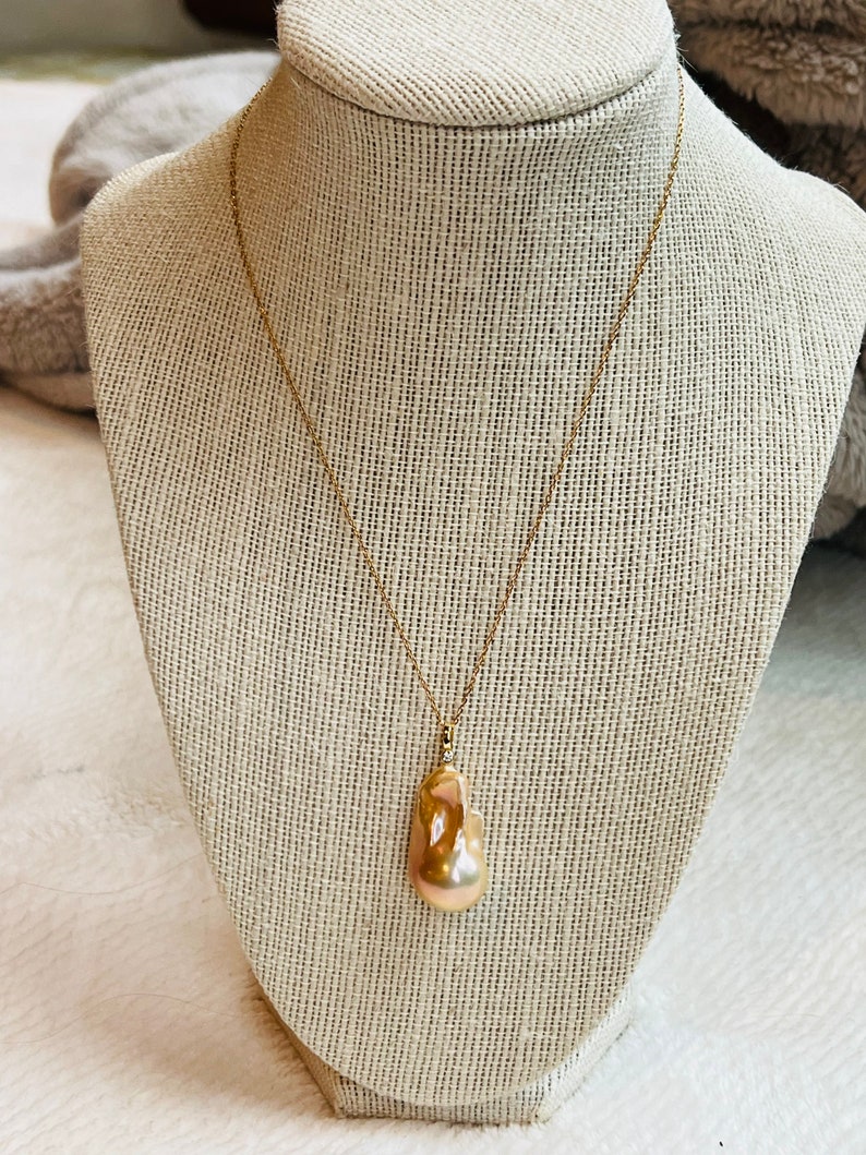 May include: A gold-colored necklace with a large, irregularly shaped, peach-colored pearl pendant. The pearl hangs from a delicate gold chain, displayed on a neutral-toned jewelry bust.