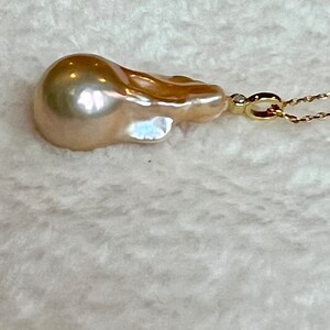 May include: A lustrous, teardrop-shaped, golden-hued pearl pendant. The pearl is attached to a delicate gold chain with a small diamond accent. The background is a soft, textured white fabric.