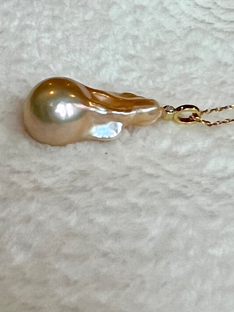 May include: A lustrous, teardrop-shaped, golden-hued pearl pendant. The pearl is attached to a delicate gold chain with a small diamond accent. The background is a soft, textured white fabric.