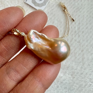 May include: A large, irregularly shaped, iridescent pearl pendant with a gold chain. The pearl has a creamy, peach color with hints of pink and gold. A small diamond accents the gold bail. The tag reads "soul star meditation".