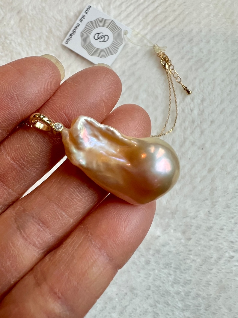 May include: A large, irregularly shaped, iridescent pearl pendant with a gold chain. The pearl has a creamy, peach color with hints of pink and gold. A small diamond accents the gold bail. The tag reads "soul star meditation".