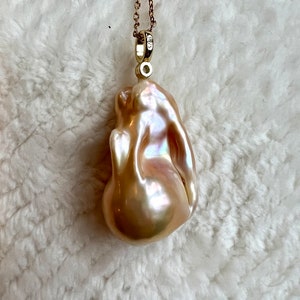 May include: A lustrous, irregularly shaped, peach-colored pearl pendant suspended from a delicate gold chain. The pearl has a smooth, iridescent surface with hints of pink and cream. The gold chain is finely linked and attached to a small gold bail.