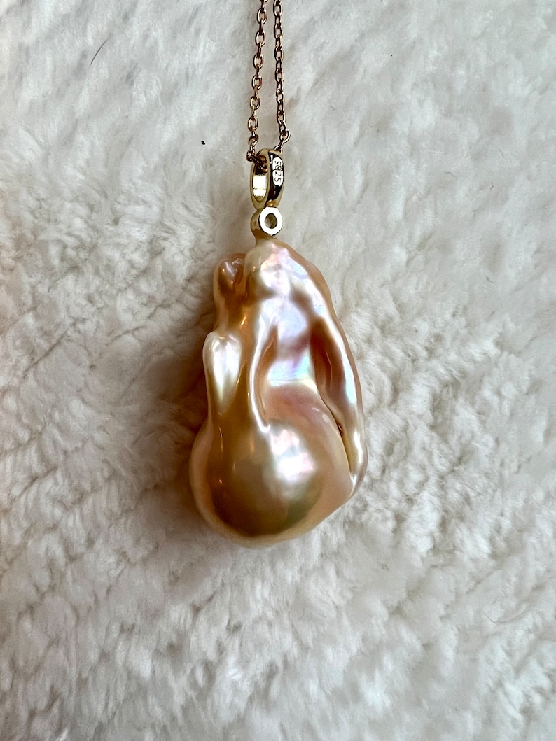 May include: A lustrous, irregularly shaped, peach-colored pearl pendant suspended from a delicate gold chain. The pearl has a smooth, iridescent surface with hints of pink and cream. The gold chain is finely linked and attached to a small gold bail.
