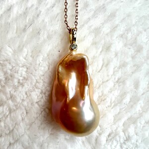 May include: A gold-toned necklace featuring a large, irregularly shaped, lustrous pearl pendant. The pearl has a warm, golden hue with iridescent highlights. A small, round diamond accents the pendant's bail. The chain is delicate and gold-colored.