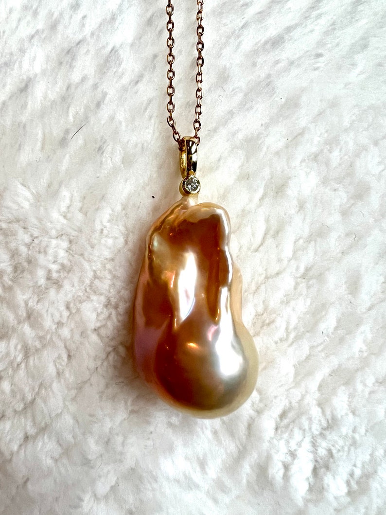 May include: A gold-toned necklace featuring a large, irregularly shaped, lustrous pearl pendant. The pearl has a warm, golden hue with iridescent highlights. A small, round diamond accents the pendant's bail. The chain is delicate and gold-colored.