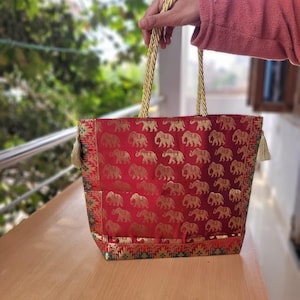 Op de afbeelding: Rode tote bag met een gouden olifantenpatroon. De tas heeft gouden touwhandvatten en kwastjes. De tas is gemaakt van een patroonstof met een rode achtergrond en gouden olifanten.