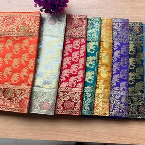May include: A selection of folded textiles in various colours, featuring gold elephant and floral patterns. The fabrics include orange, silver, red, teal, gold, purple, black, blue, and brown. These textiles are likely used for decorative purposes.