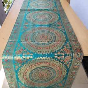 Op de afbeelding: Teal tafelloper met ingewikkelde gouden en rode cirkelpatronen. De loper heeft een gedetailleerde bloemenrand en decoratieve kwastjes aan elk uiteinde. Hij ligt op een lichtgekleurde houten tafel.