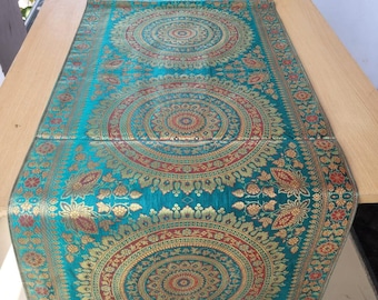 May include: Teal table runner with intricate gold and red circular patterns. The runner features a detailed floral border and decorative tassels at each end. The runner is placed on a light-colored wooden table.