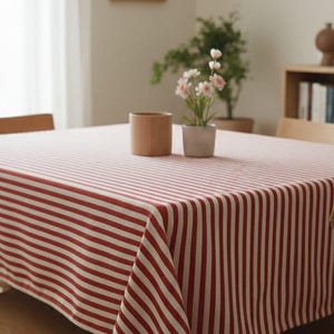 Può includere: Una tovaglia a righe rosse e bianche copre un tavolo di legno. La tovaglia è realizzata in tessuto e presenta strisce verticali. Una piccola pianta in vaso e una tazza di ceramica marrone sono sul tavolo.