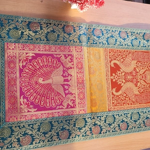 May include: A colorful table runner with a peacock and floral design. The runner features a teal border with gold accents, and panels of pink, yellow, orange, and blue. The peacock and floral patterns are in gold.