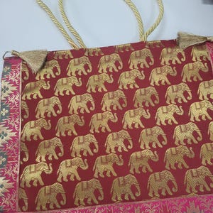 Op de afbeelding: Een rode draagtas met een herhalend gouden olifantenpatroon. De tas heeft gouden touwhandvatten en decoratieve gouden kwastjes. De randen zijn afgewerkt met een roze en gouden patroonstof.