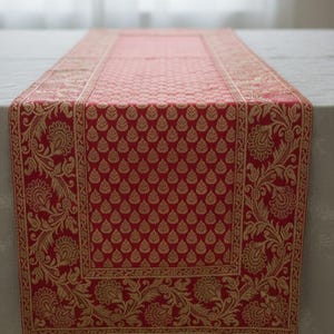 Puede incluir: Camino de mesa rojo con un patrón de hojas doradas y un borde floral dorado ornamentado. El camino de mesa rectangular está hecho de una tela texturizada, agregando un toque de elegancia a una mesa.