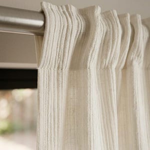 Pode incluir: Close-up de um painel de cortina creme pendurado em uma barra de prata. A cortina tem um padrão listrado vertical texturizado. O tecido é espesso e opaco, proporcionando privacidade e controle de luz. A cortina é provavelmente feita de linho ou fibra natural semelhante.