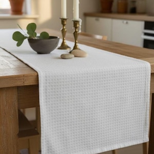 Puede incluir: Un camino de mesa blanco con tejido de gofre sobre una mesa de madera. El camino de mesa es rectangular y cubre el centro de la mesa. Dos candelabros de latón con velas blancas, un cuenco gris con vegetación y dos piedras están sobre la mesa.