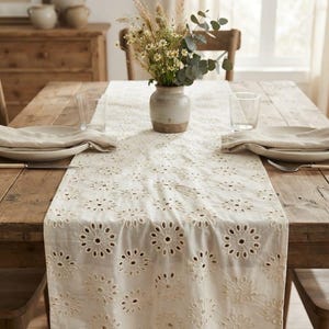 Può includere: Un runner da tavola color crema caratterizzato da un intricato motivo a ricamo inglese con margherite ripetute. Il tessuto è drappeggiato su un tavolo da pranzo in legno rustico, sotto un vaso in ceramica riempito di fiori di campo essiccati.