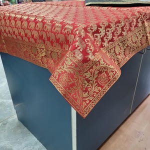 Pode incluir: Uma toalha de mesa vermelha com um padrão de elefantes dourados e uma borda dourada. A toalha de mesa está drapeada sobre um armário azul. O padrão de elefantes se repete no tecido, criando um design ornamentado e decorativo.
