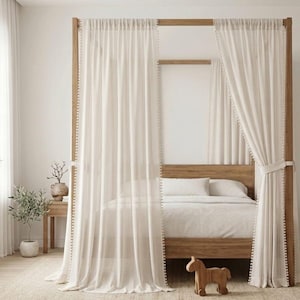 May include: A four-poster wooden bed frame draped with sheer, off-white vertical striped curtains featuring pom-pom trim along the edges. The curtains are tied back on one side, revealing a bed with white bedding and pillows within the frame.