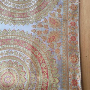 Op de afbeelding: Close-up van een decoratief textiel met een herhalend cirkelvormig patroon. Het ontwerp bevat gouden, rode en lichtgroene bloemen en geometrische elementen op een zilveren achtergrond. De stof heeft een glanzende, getextureerde uitstraling.