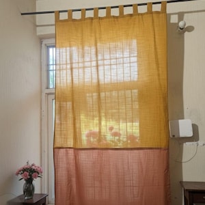 Puede incluir: Una cortina bicolor en amarillo mostaza y melocotón cuelga de una barra negra. La sección superior es de tejido texturizado y semitransparente, mientras que la inferior es de color melocotón liso. La cortina está colgada frente a una ventana.