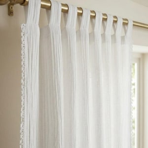 May include: White, semi-sheer curtain panels hanging from a gold-colored rod. The curtains have a textured appearance and are attached to the rod with fabric loops. The edge of the curtain has a delicate, decorative trim.