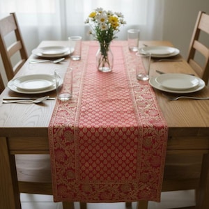 May include: A wooden dining table set for a meal, featuring a red and gold patterned table runner. White plates, clear glasses, and silverware are arranged for each place setting. A vase of flowers sits in the center.