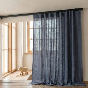 May include: A blue linen curtain hangs from a black rod, partially covering a window. The curtain has a textured appearance and is slightly translucent, allowing light to filter through. The window frame is visible behind the curtain.
