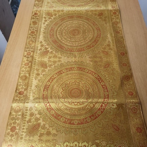 Op de afbeelding: Goudkleurige tafelloper met een herhalend cirkelvormig patroon in rood en goud. De loper heeft een gedetailleerde bloemenrand en wordt gepresenteerd op een houten tafel. Het ontwerp is symmetrisch en sierlijk.