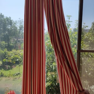 May include: Red and white striped curtains hanging in front of a window. The curtains are made of a fabric with vertical stripes. The window looks out onto a sunny outdoor scene with green trees and foliage.