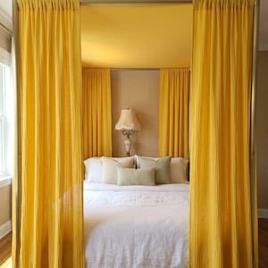 May include: A four-poster bed frame draped with vibrant yellow curtains that hang from the top rails and enclose the sides. The bed features a white duvet cover and several light-coloured throw pillows, with a lamp visible in the background.