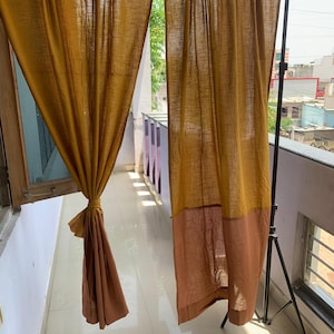 May include: Mustard yellow and brown linen curtains hanging on a black metal rod. The curtains are tied back on one side, revealing a glimpse of the outside. The fabric has a textured appearance.