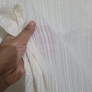 May include: Close-up of a cream-colored fabric with a vertical striped pattern. The fabric is held in a hand, showing its texture and drape. The material appears lightweight and slightly sheer.