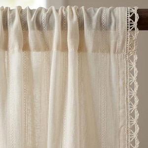 May include: Cream-colored curtain panel with a textured, semi-sheer fabric. The curtain features vertical stripes and decorative lace trim along the edge. The top is gathered, creating soft folds. The curtain is hanging on a wooden rod.