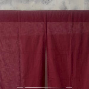 May include: Two burgundy-colored curtains hanging from a black rod. The curtains are made of a soft, flowing fabric. The top portion of the curtains has a darker, thicker band. The background is a light gray wall.