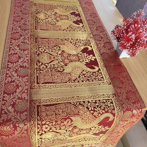 May include: A red and gold table runner with a detailed peacock design. The runner features a repeating pattern of peacocks and floral motifs, with gold accents on a red background. The edges are trimmed with a gold border and tassels.
