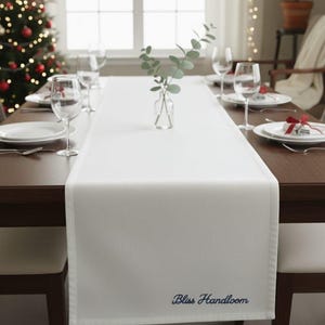 Puede incluir: Un camino de mesa blanco con el texto bordado "Bliss Handloom" en azul marino. El camino de mesa está colocado sobre una mesa de madera marrón oscuro preparada para una comida, con platos, vasos y cubiertos. Un árbol de Navidad es visible en el fondo.