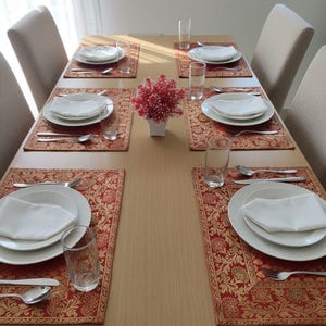 Puede incluir: Una mesa de comedor para seis personas con platos blancos, servilletas y cubiertos sobre manteles individuales rojos y dorados ornamentados. Vasos transparentes y un pequeño arreglo floral rojo completan el ambiente. Sillas beige rodean la mesa.
