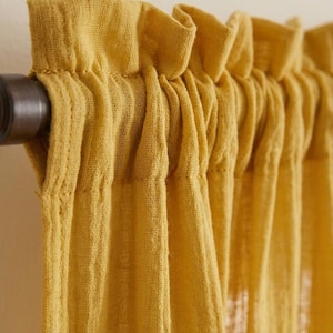 Puede incluir: Primer plano de cortinas de lino amarillo mostaza. Las cortinas cuelgan de una barra de metal oscuro. La parte superior de las cortinas tiene un diseño con volantes. La tela tiene una apariencia texturizada.