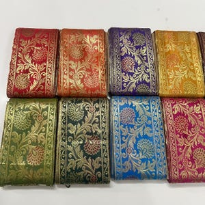 May include: A selection of decorative fabric ribbons in various colours, including red, orange, purple, yellow, green, dark green, blue, and pink. Each ribbon features a gold floral pattern.