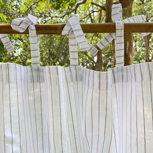 Puede incluir: Cortina blanca con rayas verticales verde oliva. La cortina se cuelga con lazos de tela blanca, anudados a una barra de madera. El fondo muestra follaje verde y ramas de árboles.